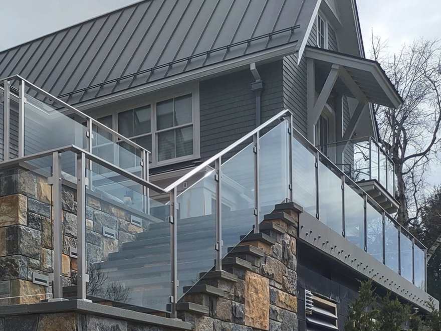 Tempered glass panels for deck railing paired with flat metal handrails create a great look. Tempered glass railing systems are custom-designed, panels are installed using glass railing mounting clamps.